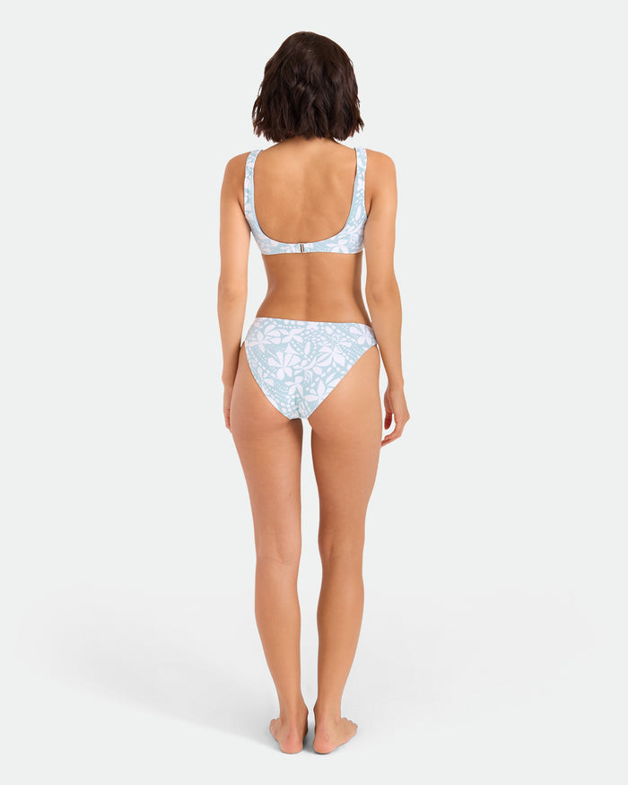 A woman stands barefoot with her back to the camera, wearing Volcom’s Women’s Tribal Stone Hipster Full Bikini Bottom in Summer Song, made from recycled nylon. The background is plain white.