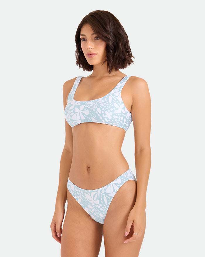 A woman with short brown hair wears Volcom's Women's Tribal Stone Hipster Full Bikini Bottom in a light blue and white pattern, standing against a plain background and looking slightly to the side.