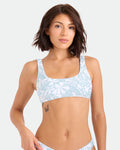 A woman with short brown hair models the Volcom Women's Tribal Stone Scoop Bralette, made from recycled nylon, in a light blue and white pattern, standing against a plain background and displaying a butterfly tattoo on her ribcage.