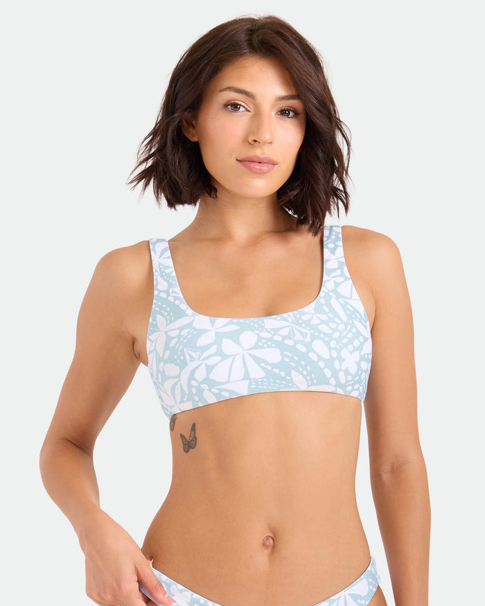 A woman with short brown hair models the Volcom Women's Tribal Stone Scoop Bralette, made from recycled nylon, in a light blue and white pattern, standing against a plain background and displaying a butterfly tattoo on her ribcage.