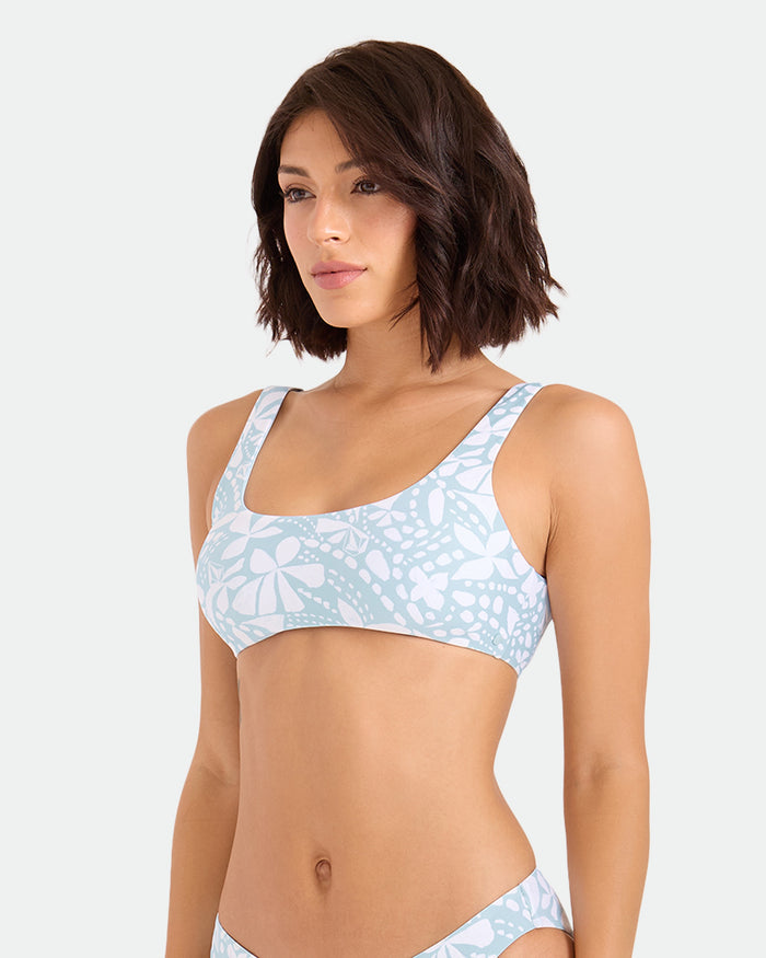 A woman with shoulder-length brown hair models the Volcom Women's Tribal Stone Scoop Bralette bikini top in a light blue and white floral print, made from recycled nylon, gazing off to the side against a plain background.