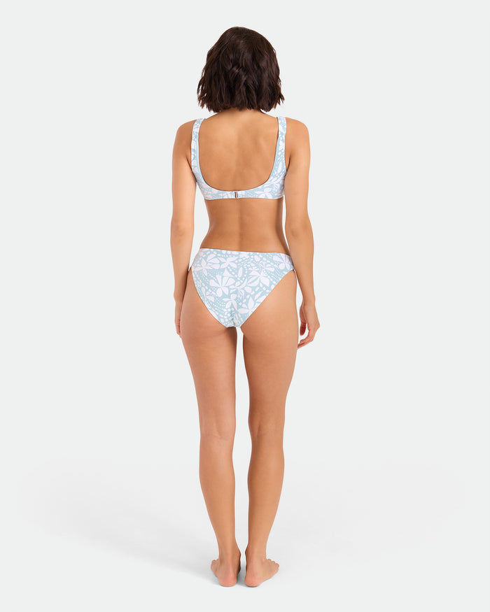 A woman stands barefoot facing away, wearing the Volcom Women's Tribal Stone Scoop Bralette bikini top in light blue and white floral recycled nylon. Her arms are relaxed at her sides, set against a plain white background.