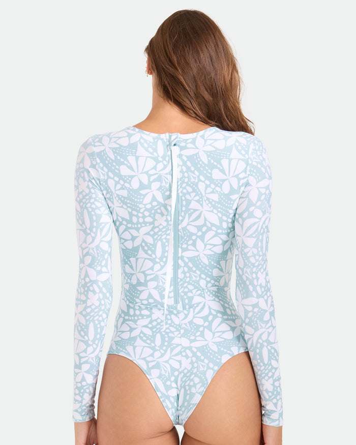 A woman with long brown hair models the Volcom Women's Tribal Stone Surf Suit, featuring a light blue and white floral print, back zipper detail, and cheeky coverage as she faces away from the camera.
