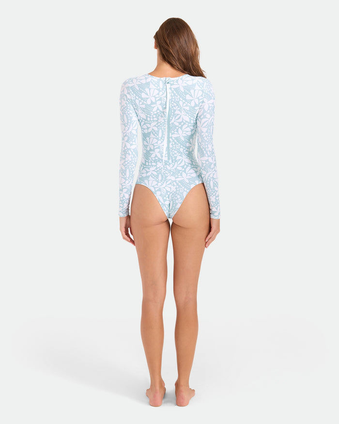 A woman stands barefoot facing away in the Volcom Women's Tribal Stone Surf Suit, a light blue and white floral long sleeve one-piece with cheeky coverage, back zipper, and recycled nylon. The plain background is light-colored.