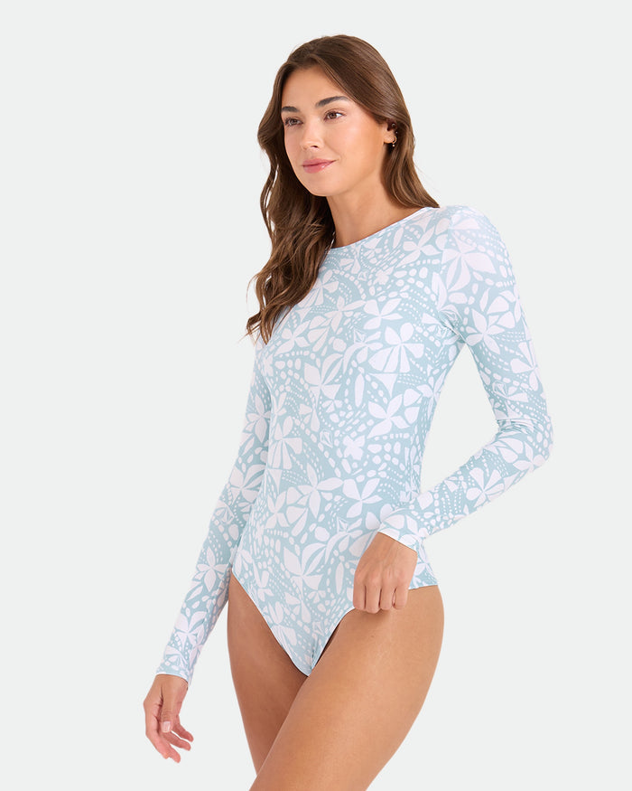 A woman models the Volcom Women's Tribal Stone Surf Suit, a long sleeve design in light blue and white floral print with cheeky coverage. She stands against a plain background, looking relaxed and gazing slightly to the side.