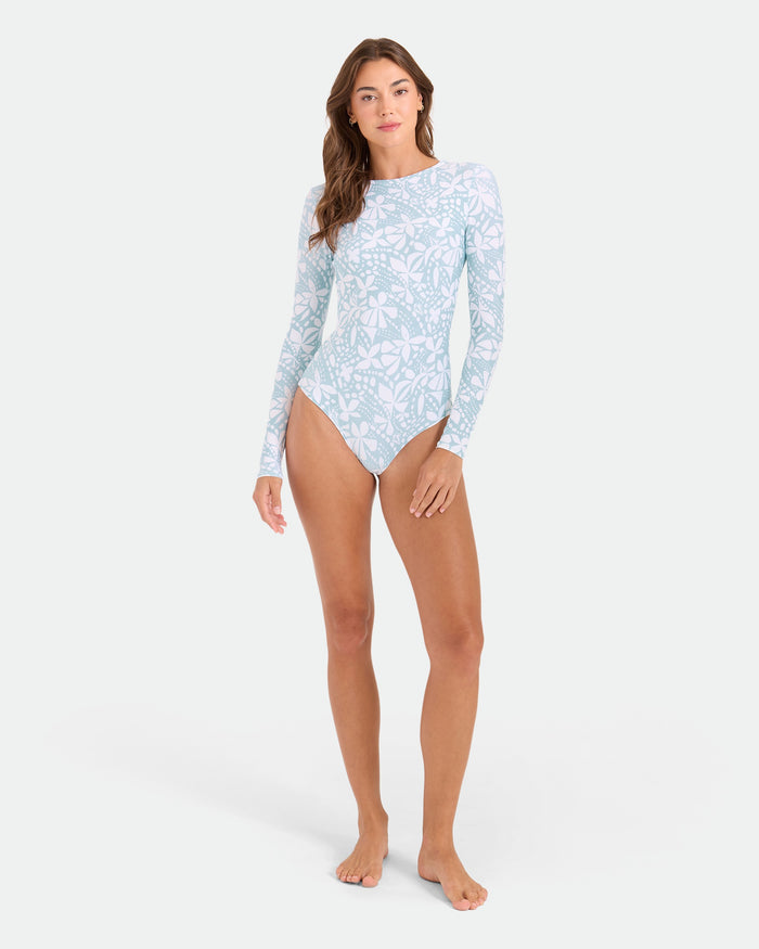 A woman stands barefoot in the Volcom Women's Tribal Stone Surf Suit, featuring long sleeves and tribal stone patterns with cheeky coverage, set against a plain white background.
