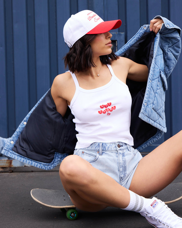 A woman wears a Volcom Women's Trippin Tank with spaghetti straps, paired with a denim jacket and shorts. She sits on a skateboard, in white-and-red cap, socks, and sneakers, holding her jacket open against a blue corrugated wall.
