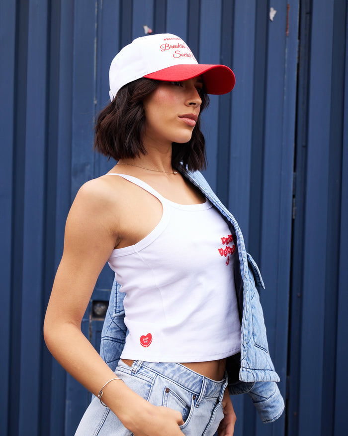 A woman in the Volcom Women's Trippin Tank with spaghetti straps, a light blue denim jacket over one shoulder, and a white and red cap stands before a dark blue corrugated metal wall, one hand in her pocket.