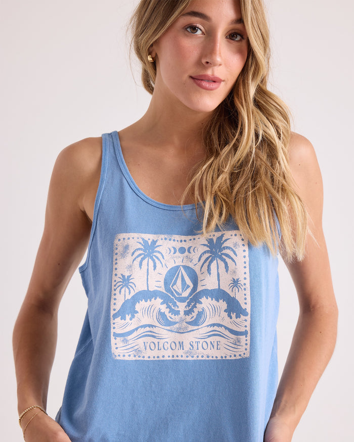 A woman with long blonde hair models the Volcom Women's Tropic Rager Bank Tank—a light blue, 100% cotton cropped tank top featuring a relaxed fit and a white Volcom Stone graphic with waves, palm trees, and the moon. She stands against a neutral background, gazing toward the camera, embodying an effortless laid-back vibe.