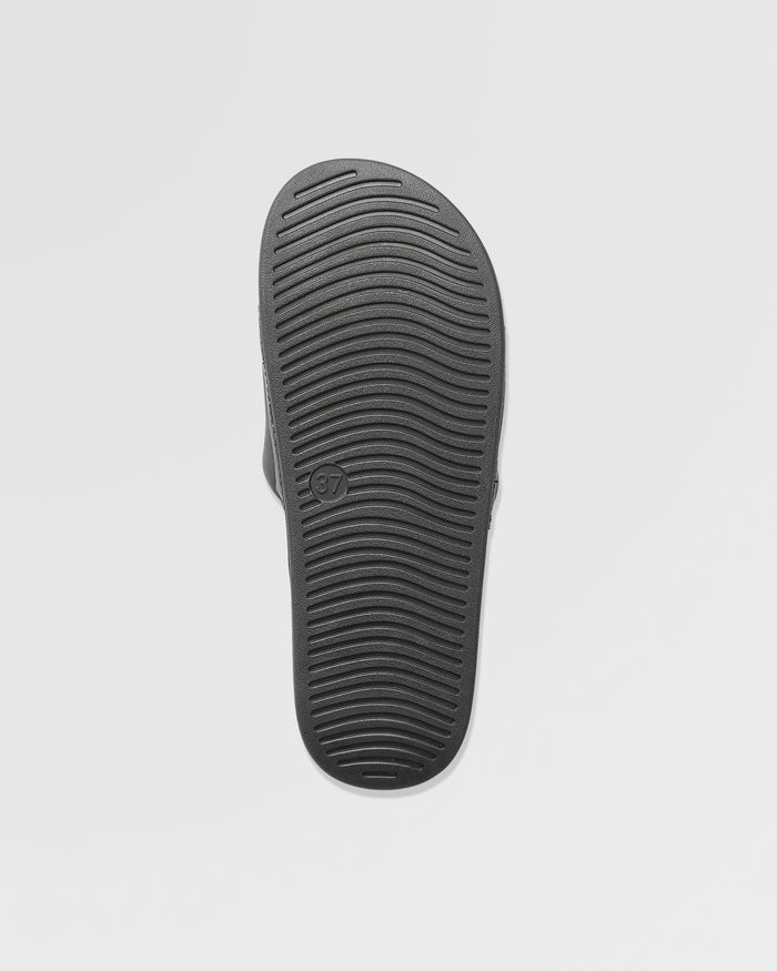 The image displays the black sole of Volcom Women's Volcation Slides, featuring horizontal grooves for traction and a size 37 mark. Set against a plain white background, it highlights everyday comfort and signature Volcom style.