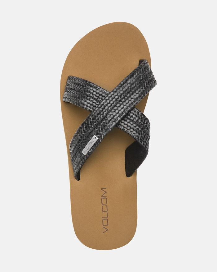 The Women's Volcom Double Cross Sandal by Volcom features a tan footbed with VOLCOM near the heel and two overlapping black textured straps forming an X across the top.