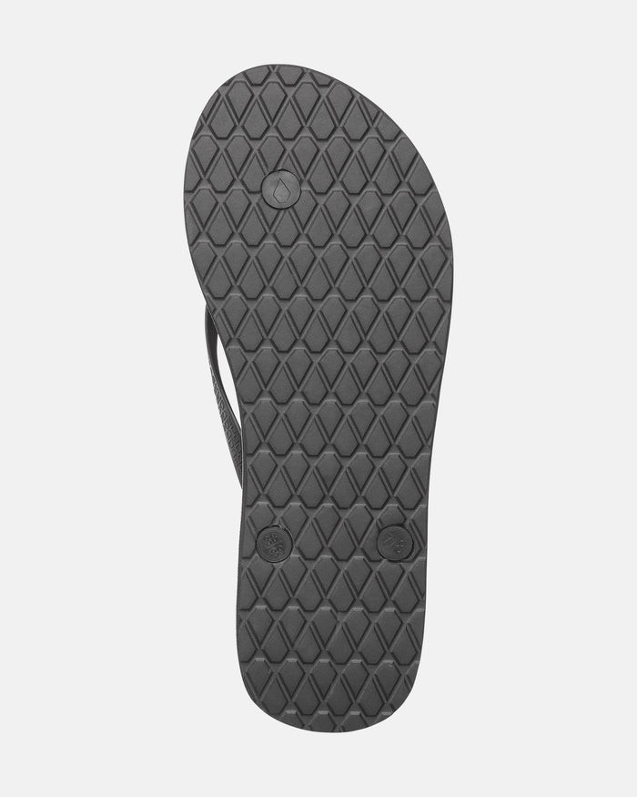 The image features the sole of the Volcom Women's Volcomotion sandal in black, displaying a diamond-patterned tread and three circular plugs underneath. Signature Volcom styling stands out against a light gray background.