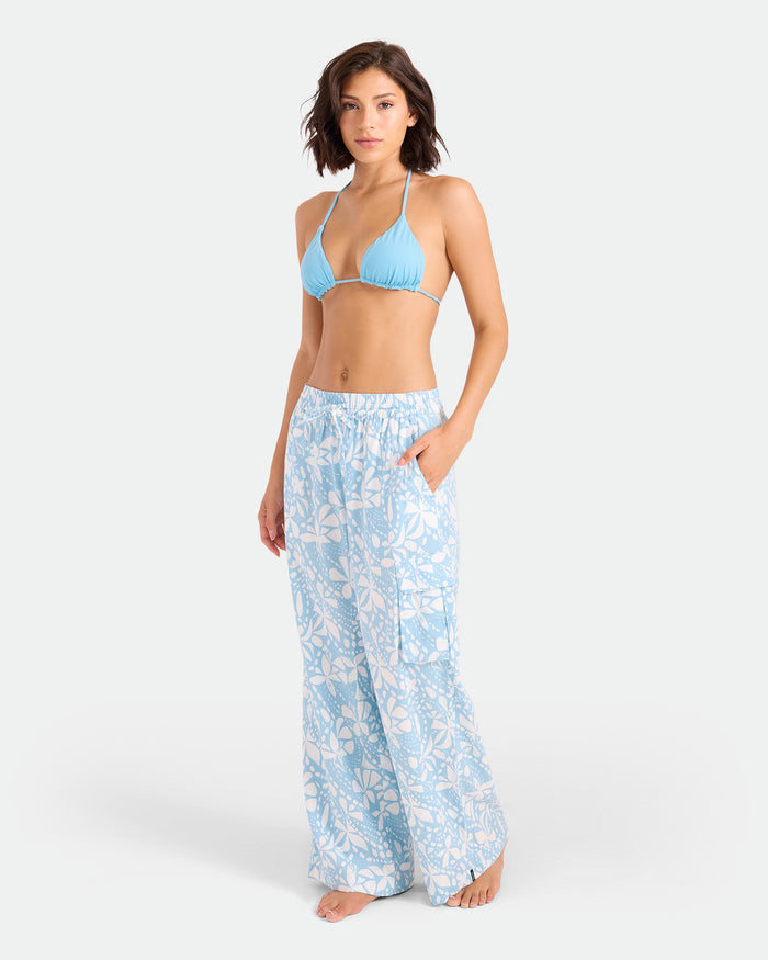 A woman stands barefoot against a plain white background, wearing a light blue bikini top and Volcom Women's Wide Leg Pull On Pants in a light blue and white floral print, featuring an elasticated waistband and her left hand in her pocket.