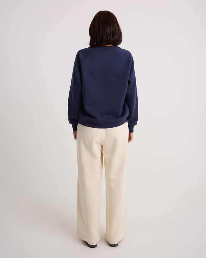 A person with shoulder-length dark hair stands facing away, wearing a navy sweatshirt and Volcom Women's Wide Leg Sweatpants in a relaxed, cream style with an elastic waistband, set against a plain, light background.