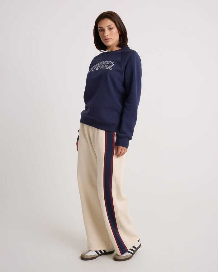A person stands against a plain background, wearing a navy sweatshirt with lettering, Volcom Women's Wide Leg Sweatpants in cream featuring an elastic waistband and red and blue side stripes, and white sneakers with dark stripes. The relaxed fit of the Volcom sweatpants enhances the laid-back look.