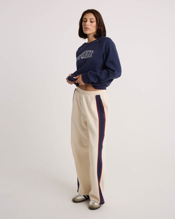 A person stands against a plain background, wearing Volcom Women's Wide Leg Sweatpants in a cream hue with navy and red side stripes. The sweatpants feature a relaxed fit and an elastic waistband. They style the look with a navy sweatshirt and black-and-white shoes, looking to the side while holding the hem of their sweatshirt.