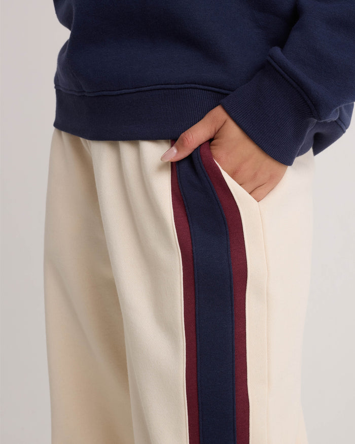 A person wearing Volcom Women's Wide Leg Sweatpants in cream, featuring navy and burgundy side stripes, a relaxed fit, and an elastic waistband, pairs them with a navy sweatshirt and places their hand in the pocket.