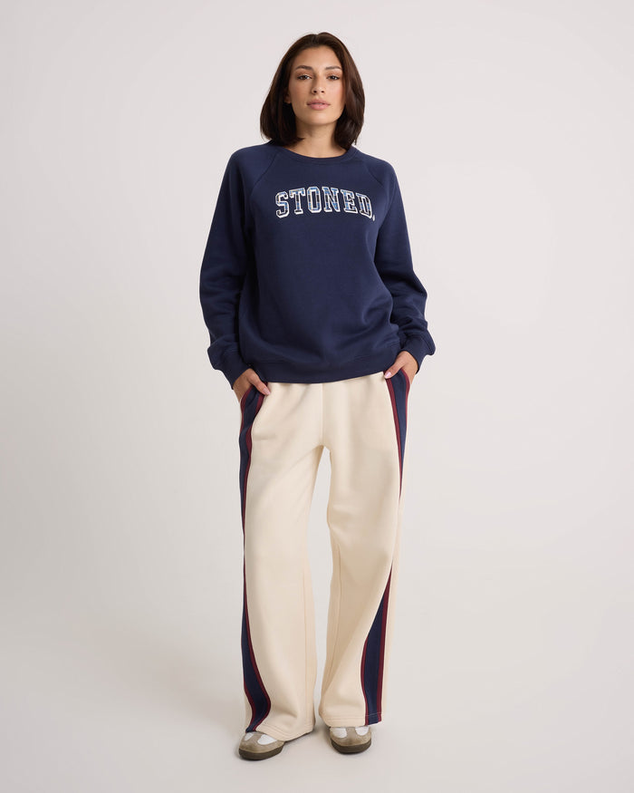 A person stands with hands in pockets, wearing a navy blue STONED sweatshirt and Volcom Women's Wide Leg Sweatpants in cream, featuring blue and red side stripes and an elastic waistband, finished with beige shoes, against a plain, light background.