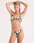A woman poses in the Volcom Women's Wild Bloom Midrise Skimpy bikini, showcasing its abstract black, white, and yellow pattern with a ribbed bottom made from recycled nylon swimwear. She stands against a plain light background with her arms raised above her head.