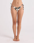 A woman stands barefoot against a plain white background, wearing the Volcom Women's Wild Bloom Tie Side Cheekini—a black, white, and yellow floral bikini bottom crafted from recycled nylon with rib texture and adjustable tie straps.