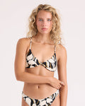 A woman with curly blonde hair wears the Volcom Women's Wild Bloom Twist Bralette bikini, which features black, white, and gold abstract shapes. She stands against a plain light background, looking directly at the camera with a neutral expression.