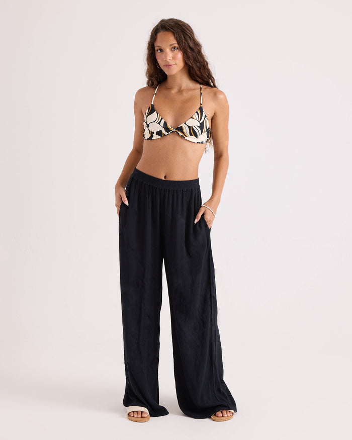 A woman with long curly hair stands against a plain background wearing the Volcom Women's Wild Bloom Twist Bralette—a patterned twist-front bikini top with adjustable straps—paired with loose black pants, sandals, and a bracelet, her hands tucked in her pockets.