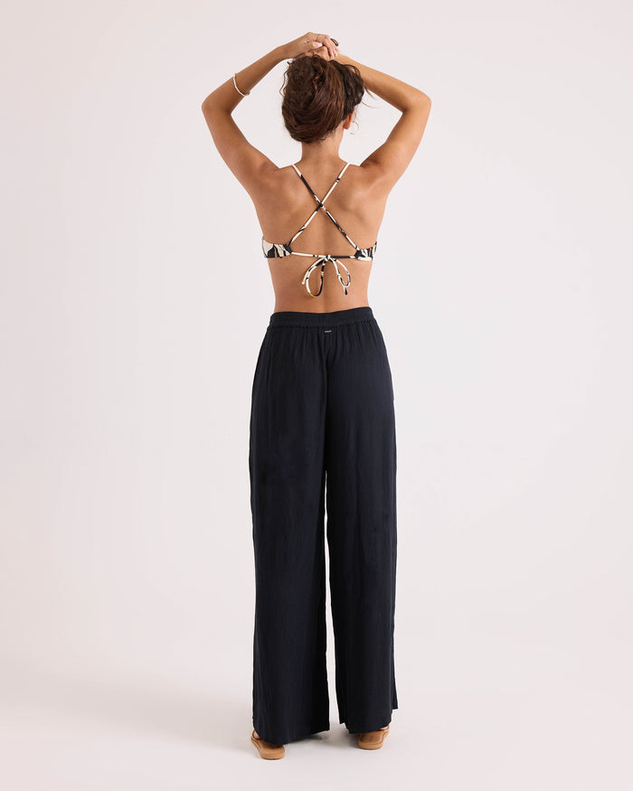A woman stands facing away, arms raised, wearing the Volcom Women's Wild Bloom Twist Bralette with adjustable straps and loose black pants against a plain light background.