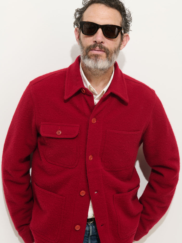 A man with short curly hair, a gray beard, and sunglasses wears the Work Jacket In Merino Wool Knit in red over a white shirt, standing against a plain white background.