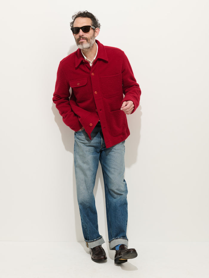 A man with dark hair and a beard, wearing sunglasses, a Work Jacket in Merino Wool Knit, blue jeans, and black shoes stands against a plain white background.