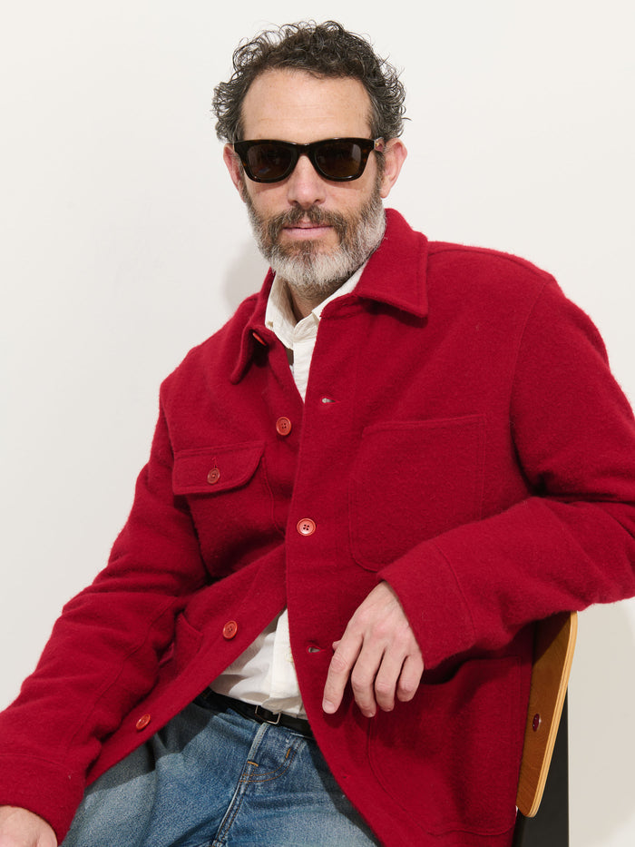 A man with curly hair, a beard, and sunglasses sits on a chair against a neutral background. He is wearing the Work Jacket In Merino Wool Knit in bright red over a white shirt and blue jeans.