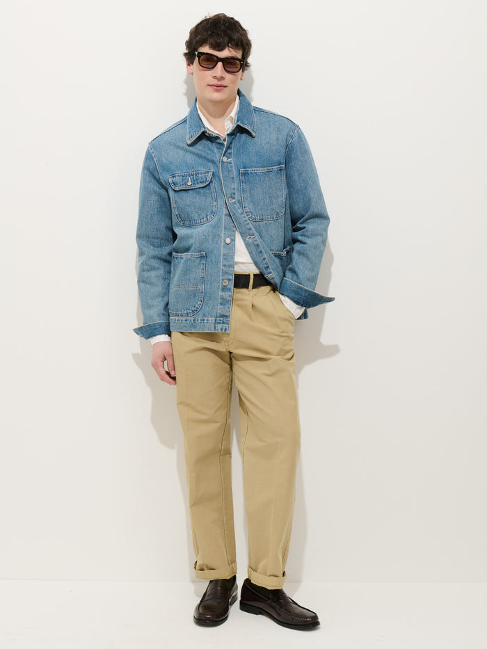 A person in dark sunglasses, a Work Jacket in Vintage Wash Denim over a white shirt, beige relaxed-fit pants, and dark shoes stands against a plain white background with one hand in their pocket.