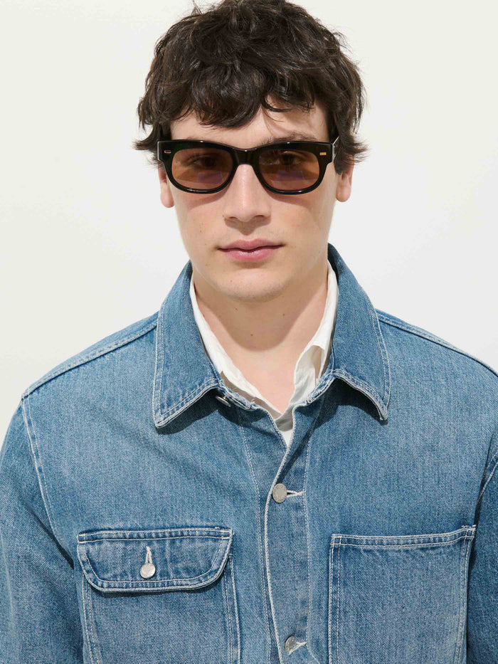 A person with short dark hair wears black sunglasses, a light shirt, and the Work Jacket in Vintage Wash Denim by Alex Mill in a relaxed fit, standing against a plain white background.