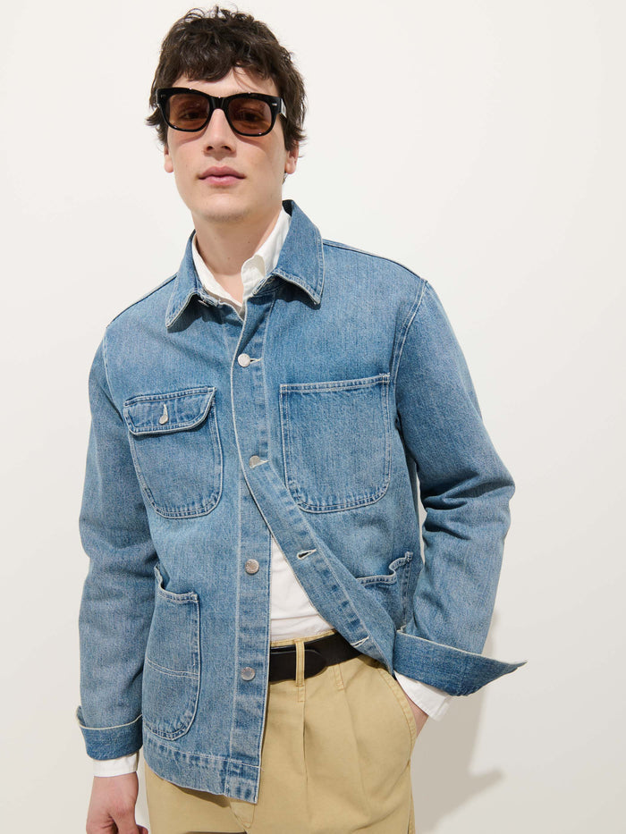 A person wears sunglasses, the Work Jacket in Vintage Wash Denim by Alex Mill over a white shirt, and relaxed fit beige pants while standing confidently against a plain white background.